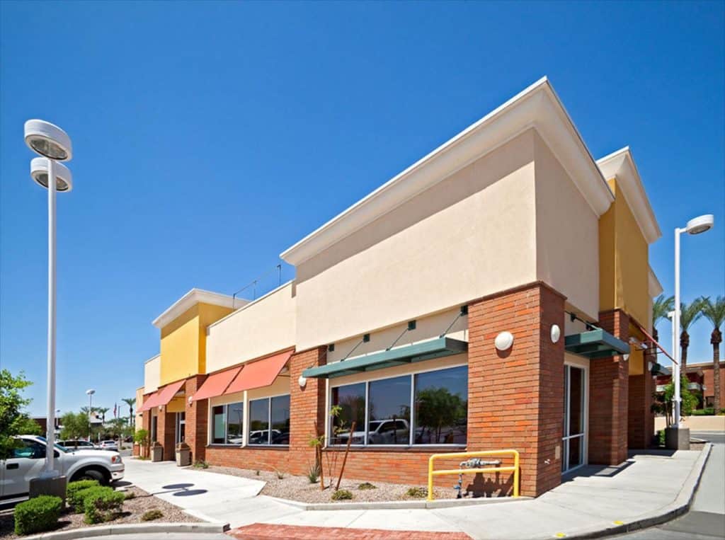 A modern, single-story commercial building with a mix of red brick and beige facade, large windows, and orange awnings gleams under the clear blue sky, thanks to the best roof washing service Northeast Kentucky offers. The scene is completed by palm trees and a convenient parking area.