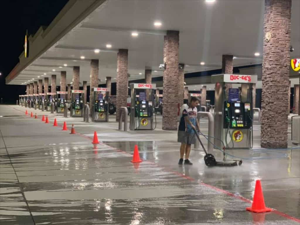 A person from a pressure washing service in Pike County, KY, is cleaning the wet concrete floor of an empty gas station at night. Several orange traffic cones are lined up along the area. The station boasts multiple fuel pumps and bright overhead lighting illuminating the scene.