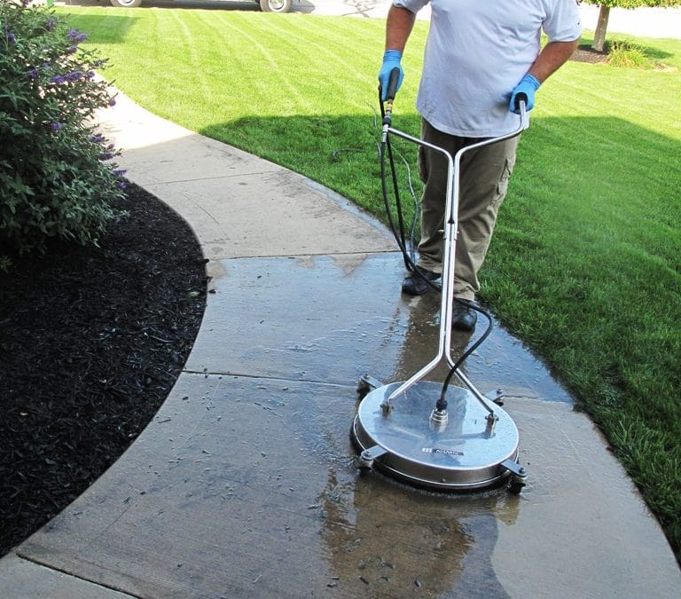 A person wearing gloves is using a circular surface cleaner attached to a pressure washer to clean a concrete pathway in Fayette County, KY. The path, set between a lawn and garden bed, sparkles wet and shiny compared to the dry concrete nearby.