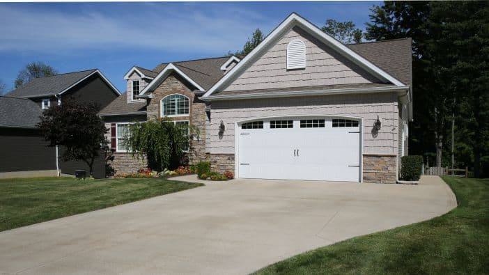 A suburban house with a two-car garage and large driveway, sparkling from a recent pressure washing service in Northeast Kentucky. The exterior features light-colored siding with stone accents. The neatly landscaped yard boasts green grass and shrubs at the front, set against a clear blue sky.
