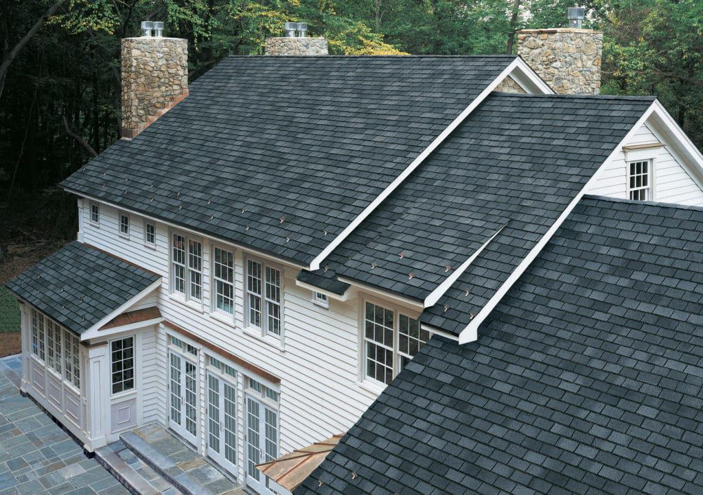 A large house with a gray shingle roof, stone chimneys, and white siding. Multiple windows sparkle thanks to the best window cleaning service in Northeast Kentucky. A paved patio completes the view as the house nestles next to a wooded area.