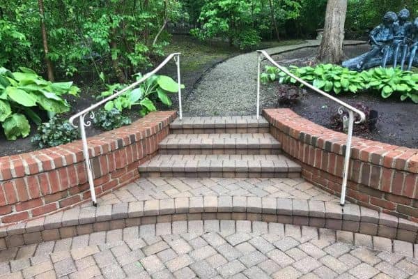 A stairway made of brick and stone pavers with four steps, flanked by metal railings, leads to a gravel path winding through a lush garden. A brick wall runs alongside the steps, which could benefit from the best pressure washing service in Pike County KY, and a sculpture peeks among the plants.