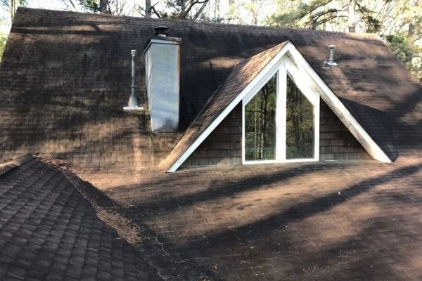 A sloped roof with brown shingles, reflecting the area's best roof washing service in Northeast Kentucky, features two chimneys and a large triangular window that mirrors the surrounding trees. Pine needles are scattered across the roof, while sunlight filters through the branches.