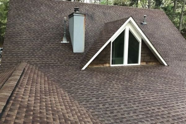 A house roof in the woods, covered in brown shingles with a steep pitch, features a prominent A-frame window and chimney. The shingles are neatly arranged, making it ideal for a pressure washing service in Pike County KY to maintain its pristine appearance.