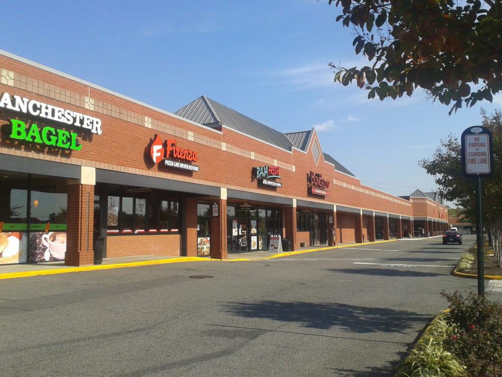 A strip mall with several storefronts, including Manchester Bagel and Frontera, showcases its red brick facade and large windows. Nearby, a "No Parking Fire Lane" sign decorates the treed parking area, kept pristine by the best roof washing service in Northeast Kentucky.