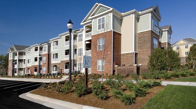 A three-story apartment building with red brick and light green siding stands out in Northeast Kentucky. It features balconies, numerous windows, and landscaped surroundings with shrubs. A lamp post and a "Speed Limit 10" sign are visible near the empty parking area, perfect for a pressure washing service.