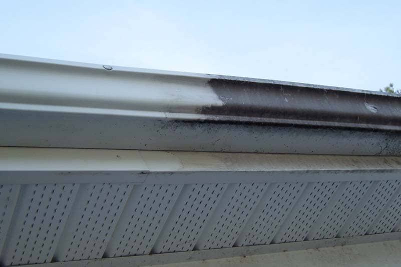 A roof gutter partially cleaned by the best roof washing service in Northeast Kentucky, revealing a stark contrast between the dirty and clean sections. The white soffit is visible beneath the gutter, set against a clear blue sky in the background.