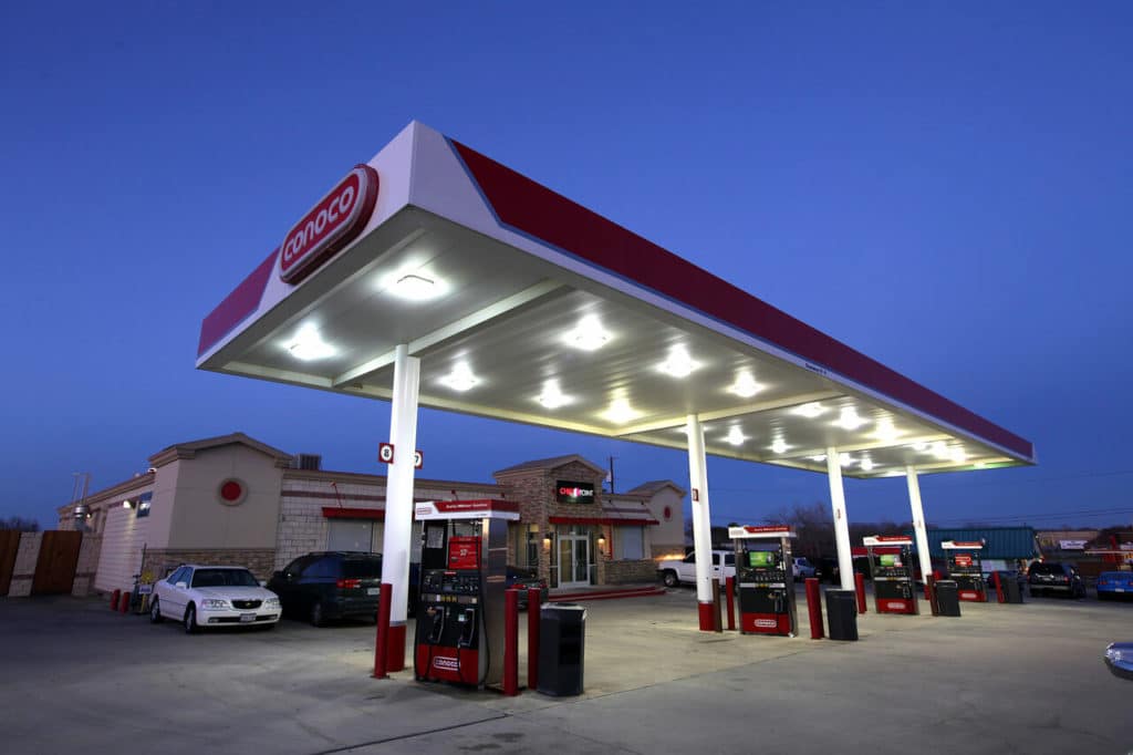 A well-lit gas station with a large canopy featuring the brand's logo and boasting the best roof washing service in Northeast Kentucky. Several spotless fuel pumps are visible under the canopy, and a few cars are parked nearby against the deep blue sky, suggesting early evening or nighttime.