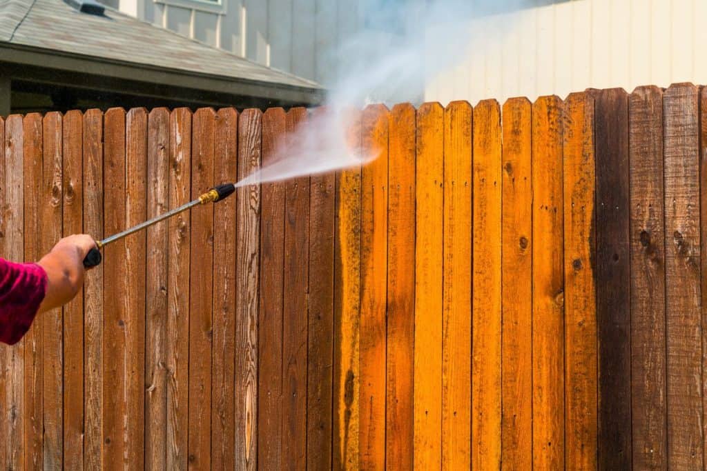 A person is power washing a wooden fence with a high-pressure spray in Fayette County, KY. The cleaned section shines brighter, free from dirt, contrasting with the darker parts. In the background, a house roof and siding accentuate the crisp results of this top-notch pressure washing service.