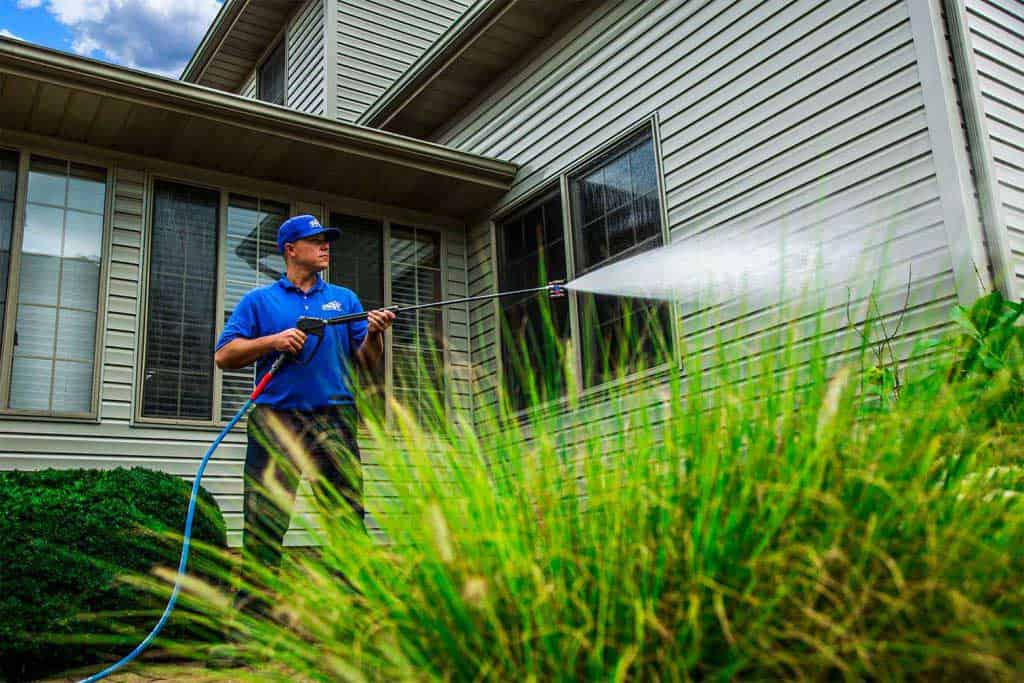 A person in a blue shirt and cap is pressure washing the beige siding of a house, framed by lush green shrubs under the bright sun. Large windows reflect the vibrant day while promoting premium pressure washing service in Fayette County, KY.