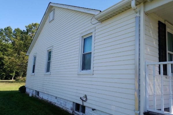 Side view of a white, single-story house with two windows, siding, and a small porch. The house sits on a grassy lawn with trees in the background under a clear blue sky, glistening as if it just received expert care from a pressure washing service in Pike County, KY.
