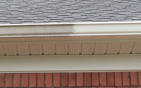 A close-up view of a house roof reveals a gutter partially cleaned, highlighting the stark contrast between clean and dirty areas. The grey shingles and red brick wall below stand out, showcasing the impressive results achievable with a pressure washing service in Cabell County, WV.