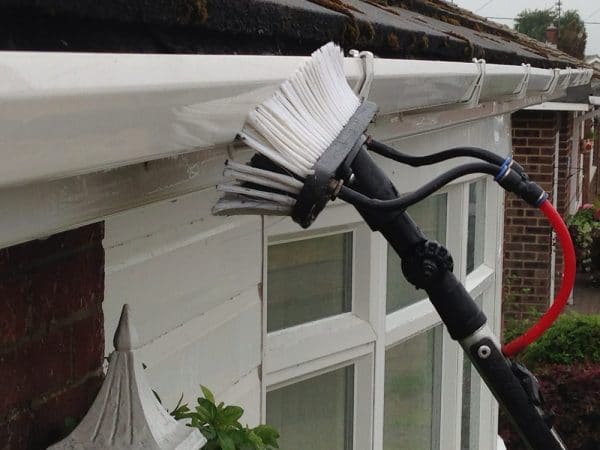 A specialized cleaning brush on a telescopic pole, part of the best roof washing service in Northeast Kentucky, is washing the outside of a gutter. It's connected to a water hose and reaches the white gutter above a window with precision and ease.