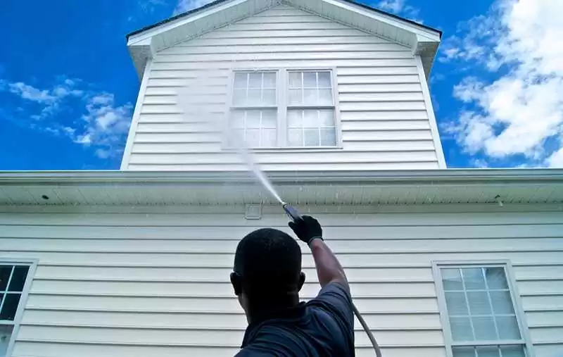 A person pressure washes the exterior of a two-story house with white siding on a sunny day. The individual, dressed in dark clothing and gloves, represents the best roof washing service in Northeast Kentucky, standing under a sky of blue with scattered clouds as they work diligently.