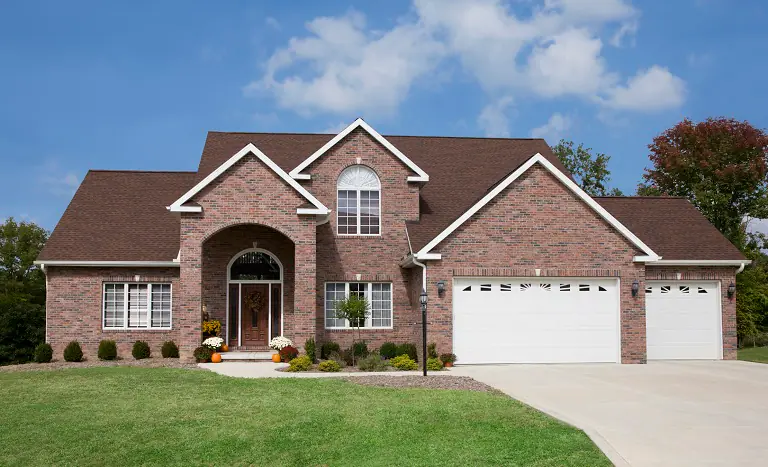 A two-story house with a red brick exterior, brown roof, and a large arched entryway gleams after the best window cleaning service in Northeast Kentucky. It features three garage doors and manicured landscaping with small shrubs. The sky is partly cloudy.