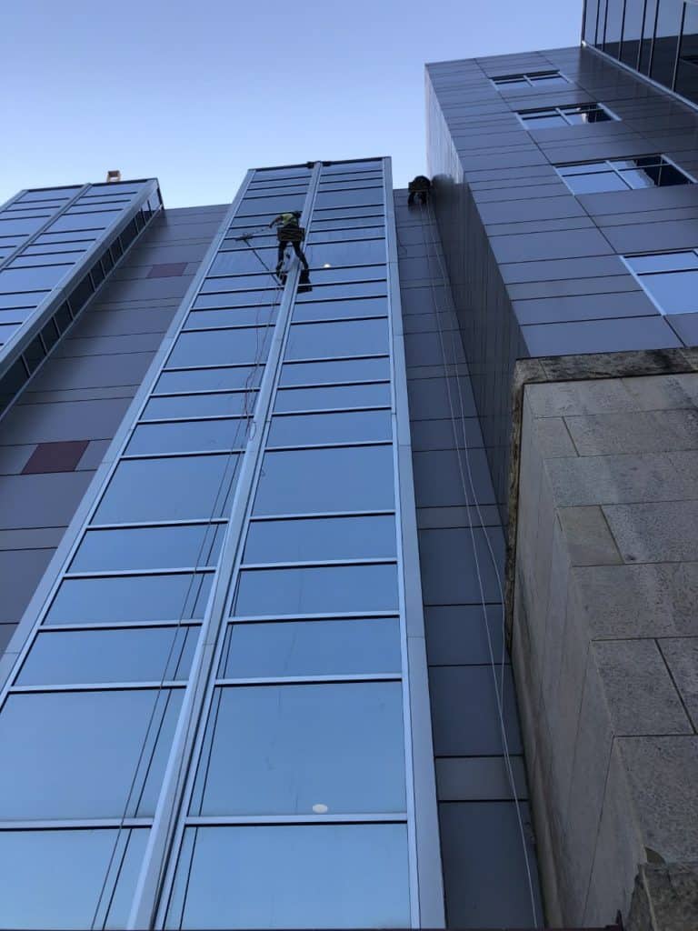 A rope access worker hangs midway up the side of a tall modern building, providing the best window cleaning service in Northeast Kentucky. Large glass windows gleam under his care, with safety ropes visible against the clear sky as they run from the worker to the building's roof.