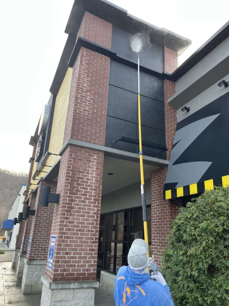 A person in a blue jacket uses a long yellow pole to pressure wash the upper part of a brick building, aiming at a black surface. This could be part of the best window cleaning service in Northeast Kentucky. The building features brick and grey walls, with a bush on one side.