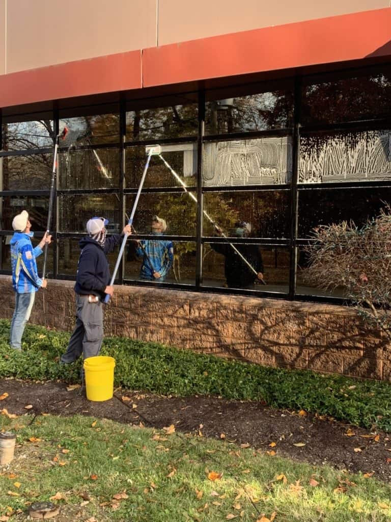Three people are cleaning large windows of a building using long-handled tools. A yellow bucket is on the ground beside them. They are outside, surrounded by grass and some bushes. The sky is clear, suggesting a sunny day—a perfect scene inspired by the best roof washing service in Northeast Kentucky.