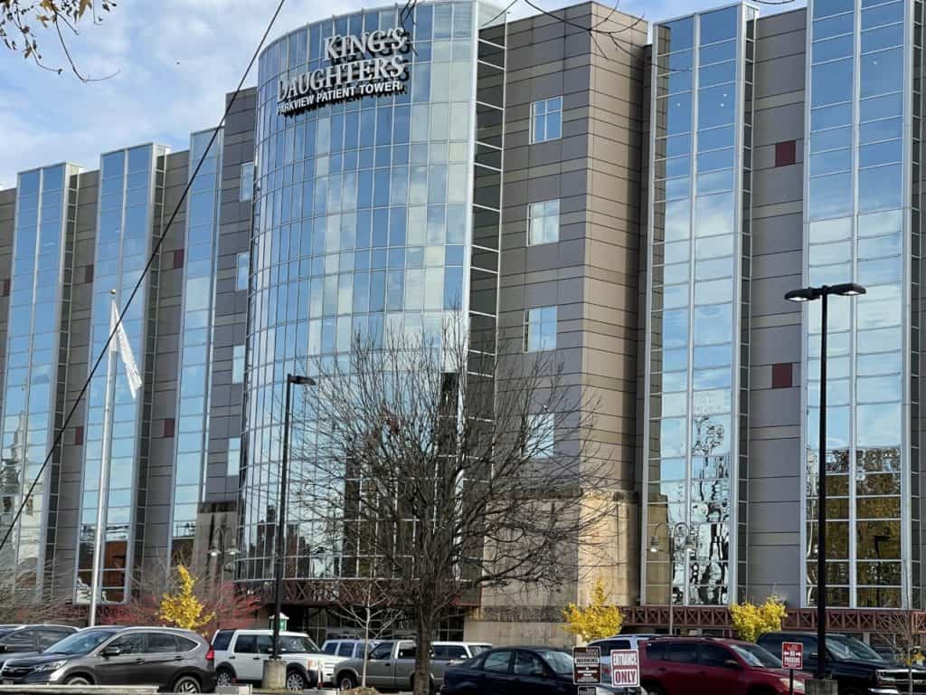 Modern hospital building with mirrored glass facade labeled "King's Daughters," featuring a central cylindrical structure. Leafless trees and parked cars are visible in the foreground, showcasing how the best roof washing service Northeast Kentucky keeps the facility's exterior pristine.
