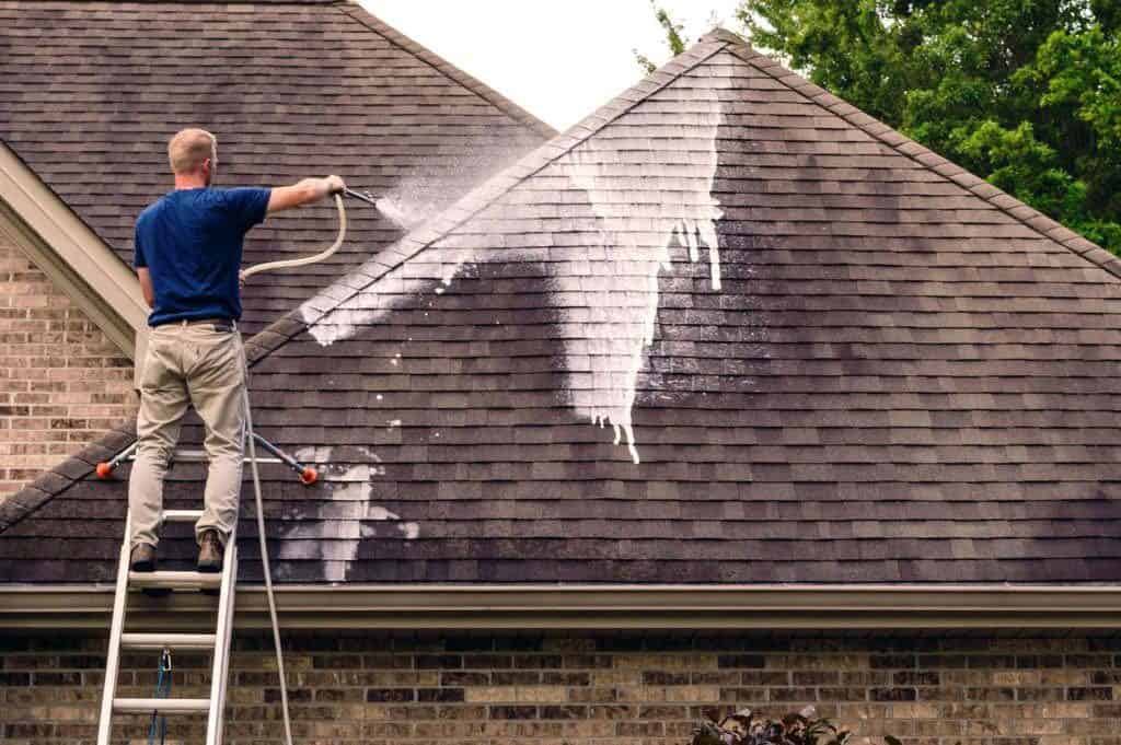 A person in a blue shirt is standing on a ladder, power washing a rooftop with dark shingles. White cleaning solution or water sprays away dirt and stains. In the background, a brick wall and green trees enhance the scene—it's an expert pressure washing service in Fayette County, KY.