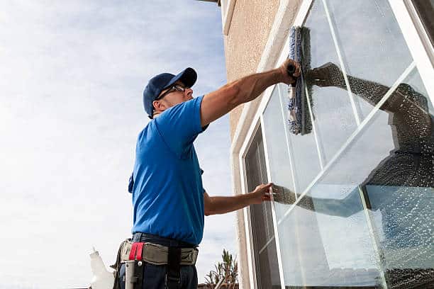 A man in a blue shirt and cap is cleaning a large window with a squeegee. Armed with cleaning supplies on his utility belt, he represents the best window cleaning service in Northeast Kentucky, ensuring crystal-clear views on this sunny day.
