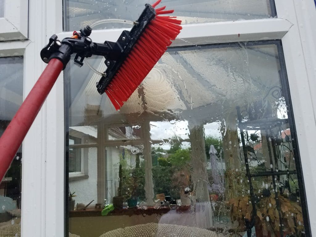 A red-bristled brush on an extendable pole is expertly cleaning a large window from the outside. Water streams down, leaving a pristine finish. Inside, a sunroom with plants and furniture is visible through the glass, highlighting why this is the best window cleaning service in Northeast Kentucky.