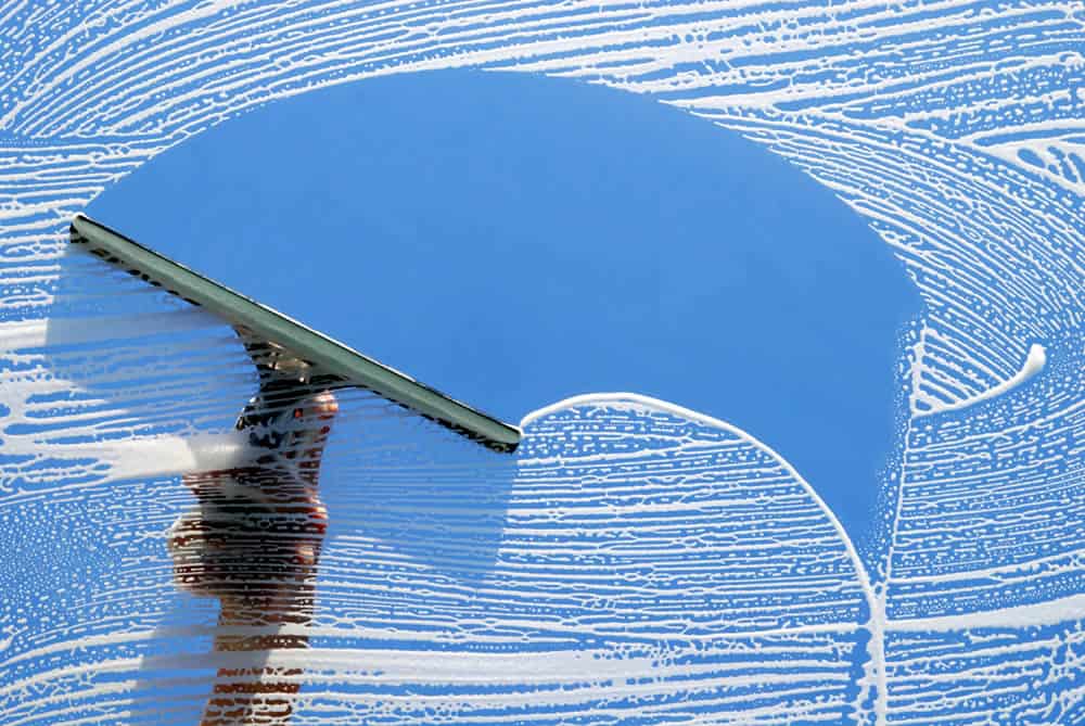 A hand using a squeegee cleans a window, leaving a clear, arch-shaped path through soapy suds. Through the cleaned section, the blue sky is visible, contrasting with the surrounding white soap—a glimpse of perfection akin to a pressure washing service in Cabell County WV.