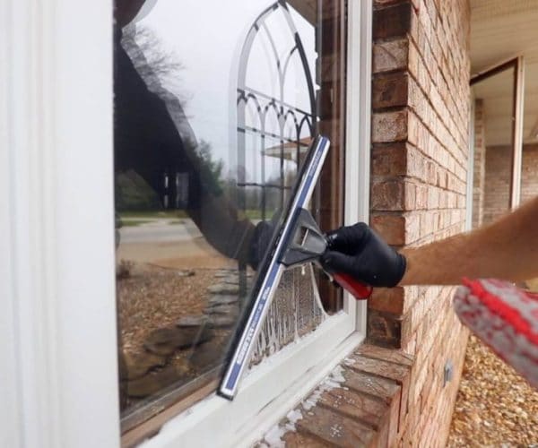 A person expertly cleans a window with a squeegee, donning a black glove and wielding a red-and-black tool. The reflection reveals trees and a driveway outside. This scene is set against a brick wall, showcasing the best window cleaning service in Northeast Kentucky.