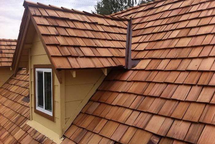 A roof with wooden shingles in a staggered pattern covers a yellow house. There is a white-framed window on the left side. The shingles vary in shades of brown, adding texture to the roof surface, ideal for maintaining through a pressure washing service in Fayette County, KY.