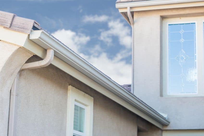 Close-up view of a house corner with a light beige stucco exterior and a gutter system along the roofline, ready for the best roof washing service in Northeast Kentucky. A decorative glass window showcases an intricate pattern against a blue sky with scattered clouds.