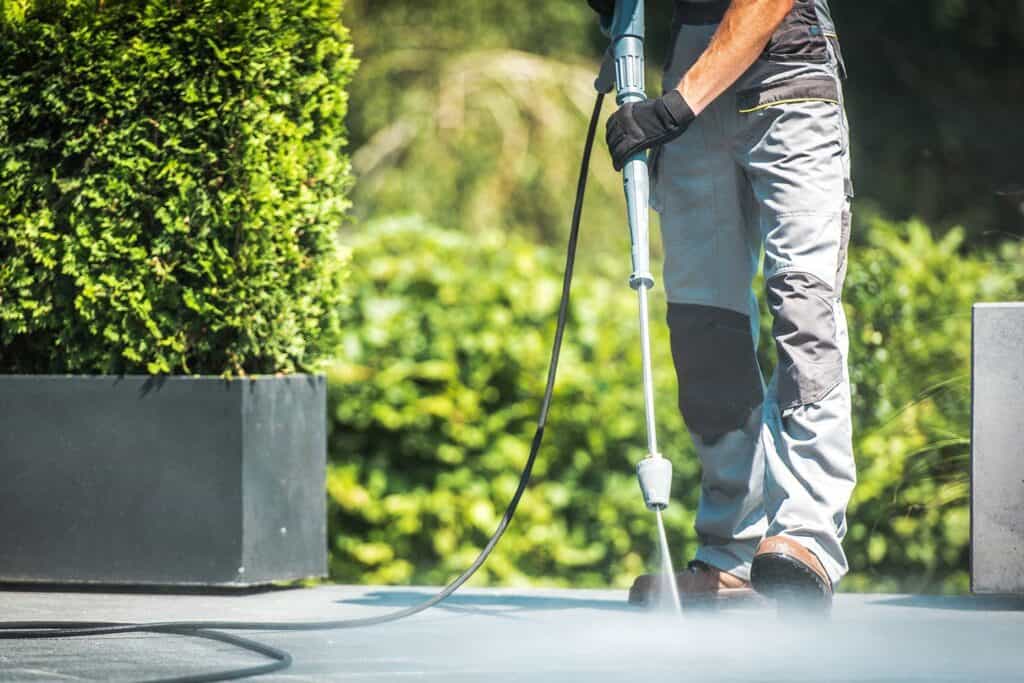 A person is pressure washing an outdoor patio, wearing gray pants, gloves, and brown shoes. Lush green plants grace the background as the powerful water jet creates a misty aura on the ground.