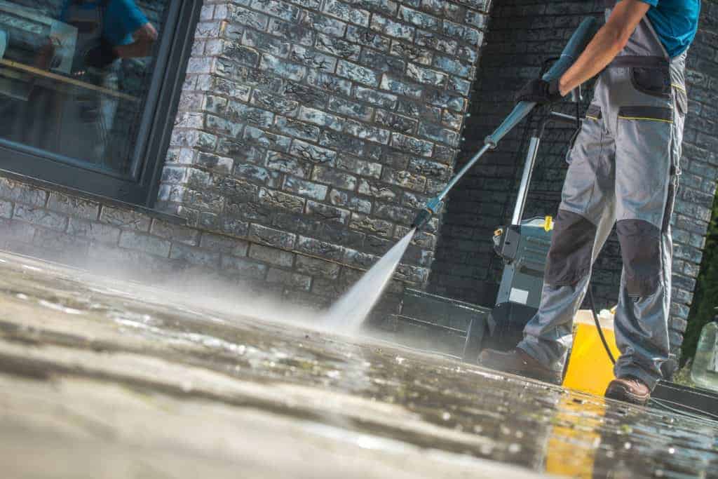 A person in work attire is pressure washing an outdoor stone patio, with a mist rising as they clean the surface. Nearby, a yellow container hints at their expertise. Behind them stands a brick building, illustrating the best roof washing service Northeast Kentucky has to offer.