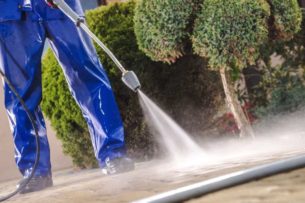 A person in blue protective clothing uses a pressure washer to clean a stone pavement, showcasing the best roof washing service in Northeast Kentucky. A green shrub adds a touch of nature to the background.