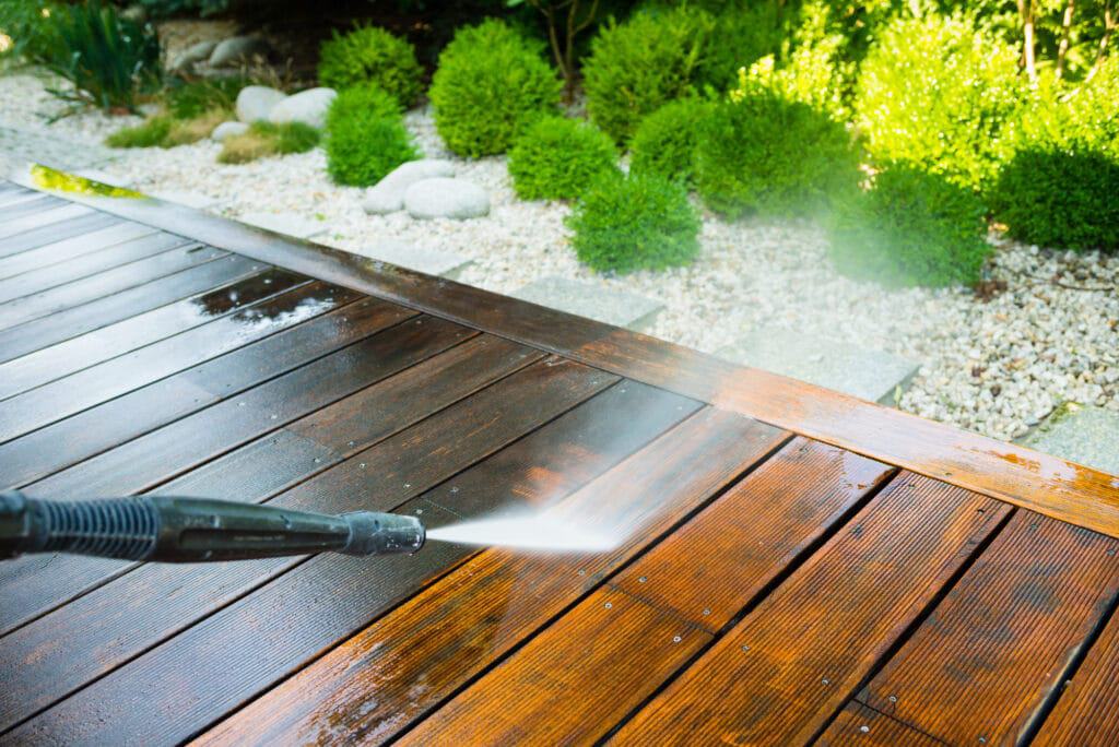 A pressure washing service in Fayette County, KY is revitalizing a wooden deck, unveiling the stark contrast between cleaned and uncleaned areas. The deck is surrounded by lush green bushes and decorative stones, creating an inviting garden setting.