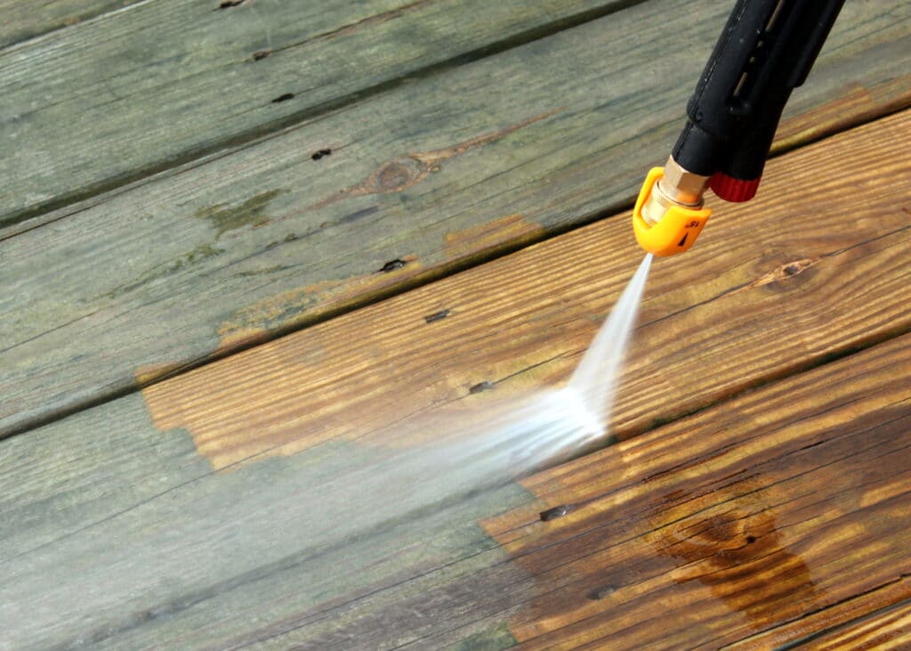Close-up of a power washing nozzle spraying water onto a wooden deck, cleaning the surface. The left side is darker and dirty, while the right is lighter and clean where the water has been applied. Experience this transformation with our pressure washing service in Cabell County, WV.