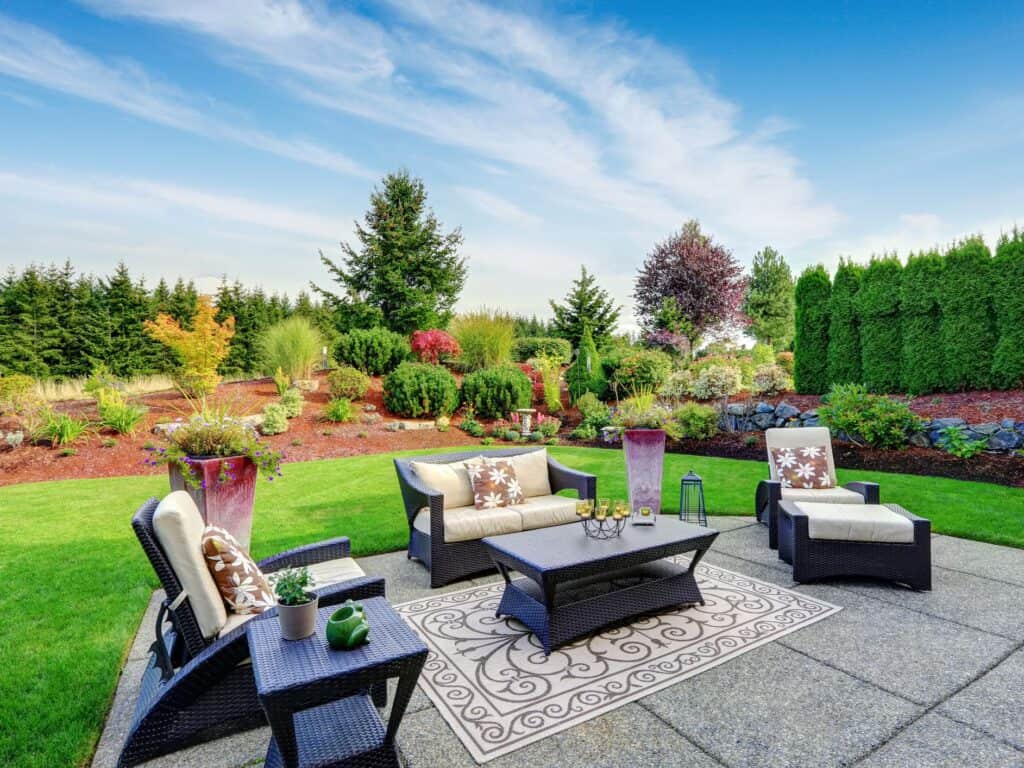 A spacious outdoor patio with wicker furniture, including a sofa, chairs, and a coffee table on a decorative rug. The lush garden features colorful flowers, shrubs, and trees under a clear blue sky—a perfect space rejuvenated by our pressure washing service in Cabell County, WV.
