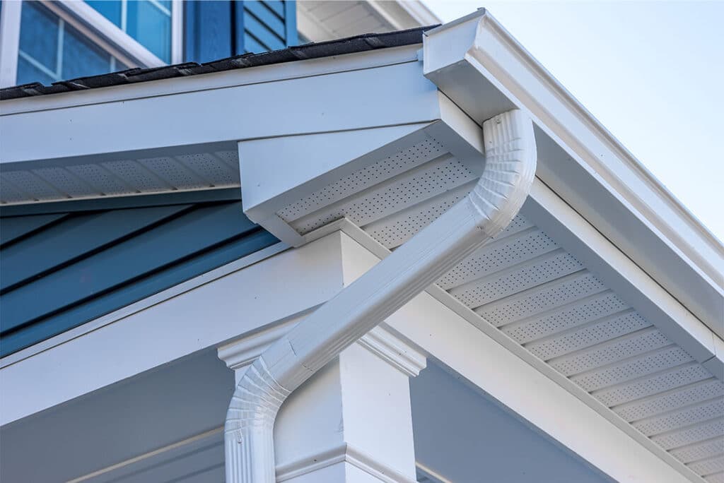 Close-up of a house's exterior showing a white gutter system attached to the roof edge, freshly cleaned by the best roof washing service in Northeast Kentucky. The downspout runs vertically along the blue and white siding, capturing architectural details under a clear sky.