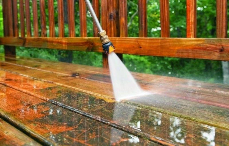 A pressure washer from the top-rated pressure washing service in Northeast Kentucky is cleaning a wooden deck. The water spray clears away dirt, revealing the natural wood color beneath. In the background, green foliage hints at an inviting outdoor setting.