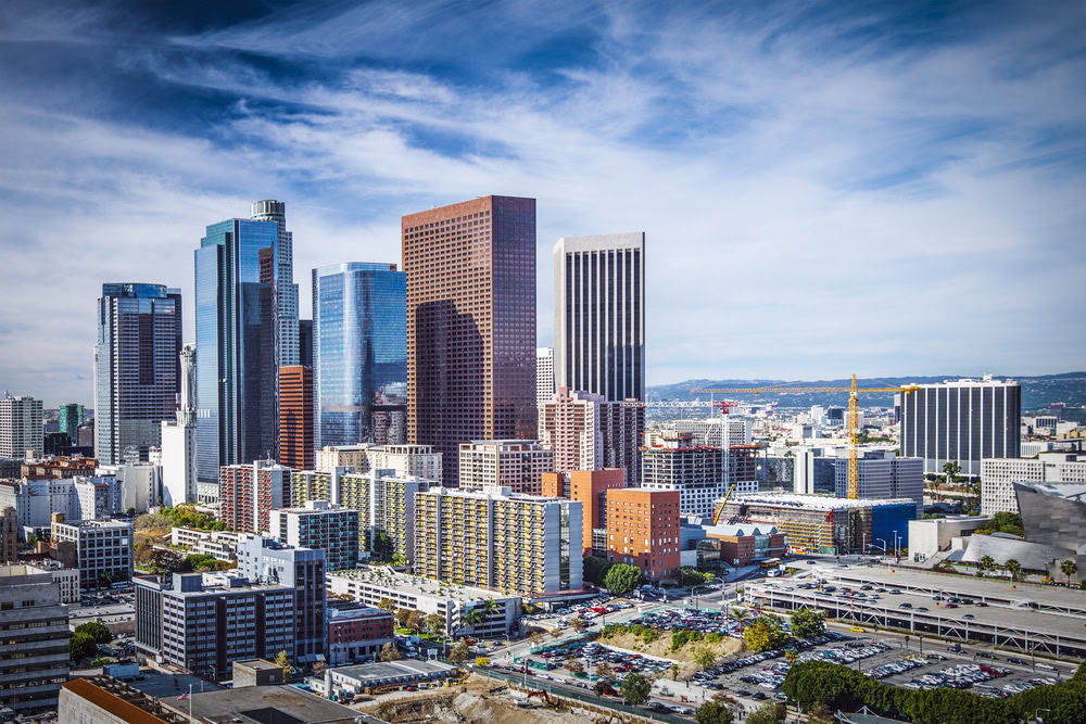 Aerial view of a city skyline with a mix of tall skyscrapers and smaller buildings under a partly cloudy blue sky. In the foreground, streets and cars bustle past parking lots where cranes hint at ongoing construction, highlighting the demand for pressure washing services in Cabell County, WV.