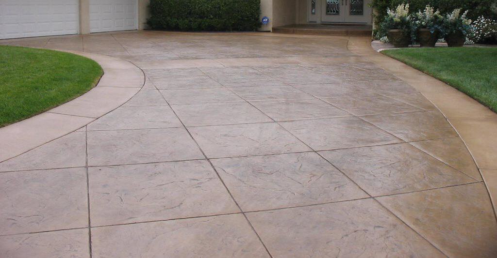 The image shows a wide, stamped concrete driveway with a subtle pattern, leading to a multi-car garage. Lush green grass borders the driveway, and potted plants are visible near the garage entrance—freshly cleaned by a pressure washing service in Fayette County, KY.