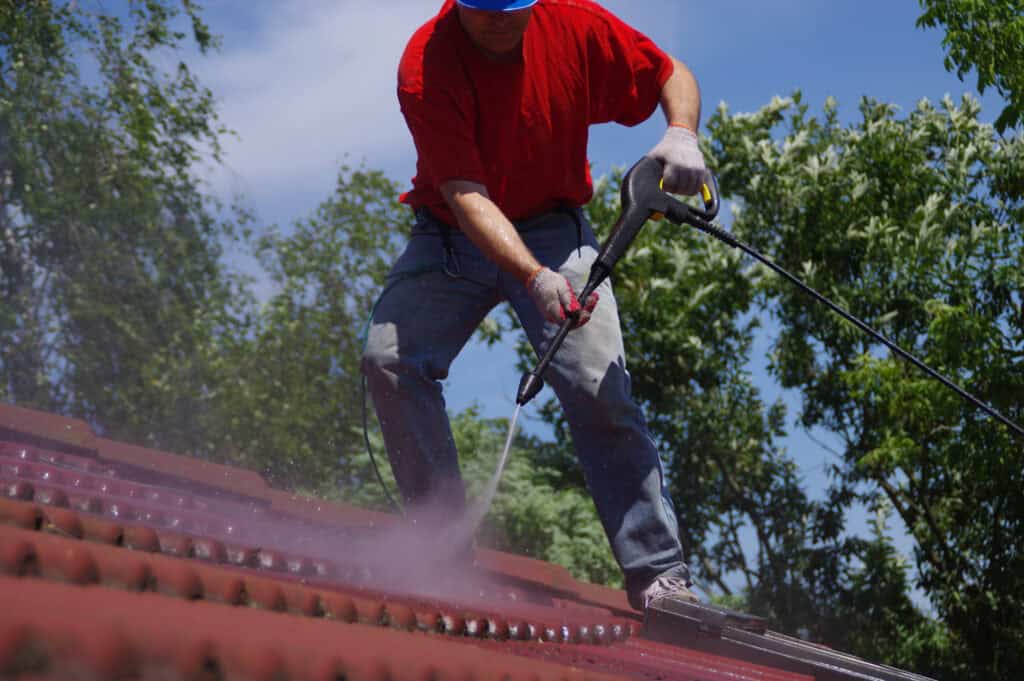 A person wearing a red shirt and blue pants uses a pressure washer to clean a red tiled roof, showcasing the best roof washing service in Northeast Kentucky. Clad in gloves and a blue hard hat, they work diligently against the backdrop of green trees and a clear blue sky.
